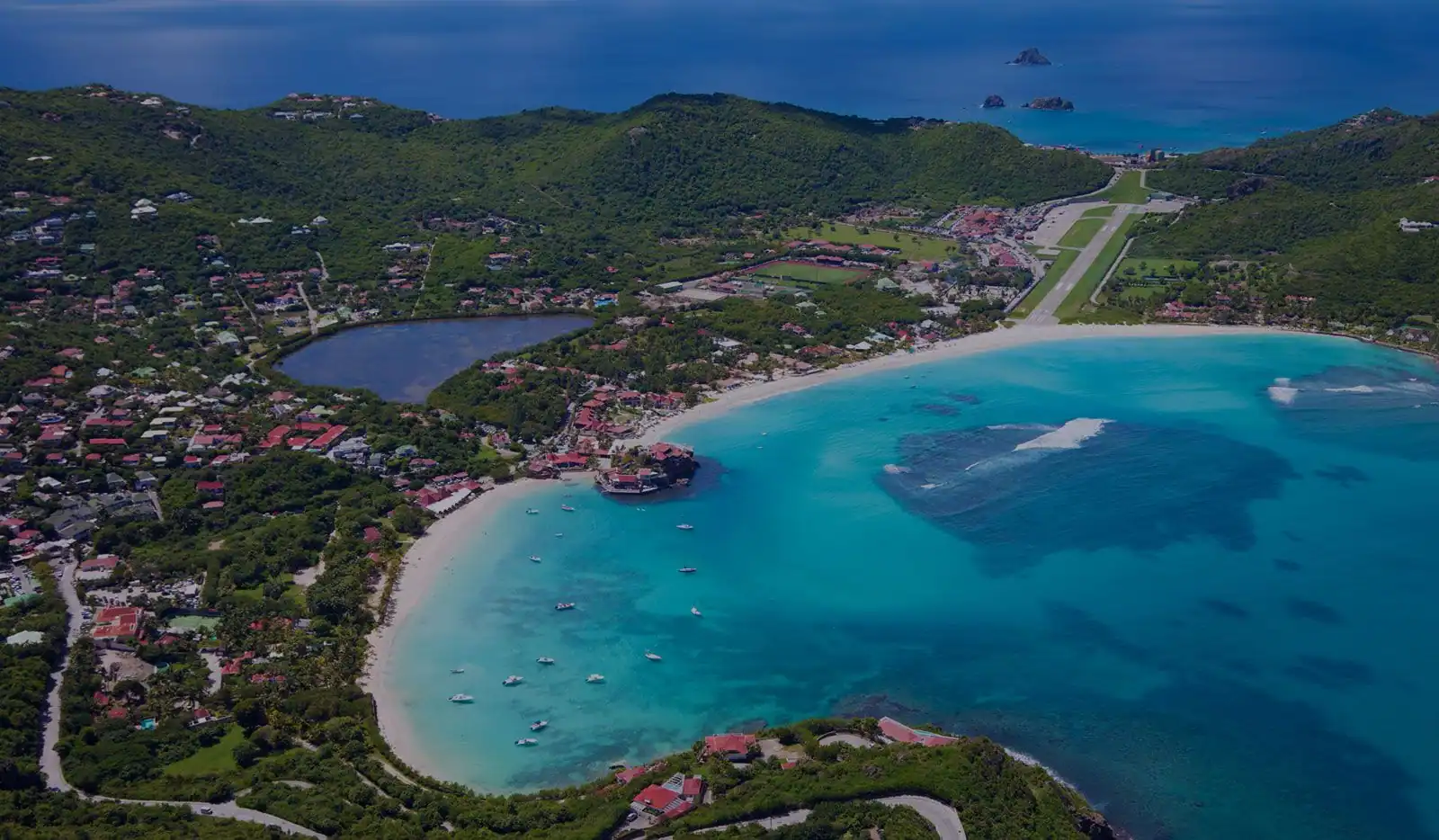 Aerial view of St. Barths island featuring turquoise bay, white sand beach, airport runway, and lush green hills