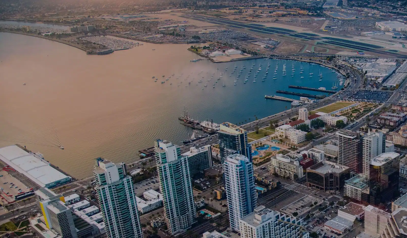 Aerial view of San Diego waterfront with high-rise buildings, marina with sailboats, and airport runway in background