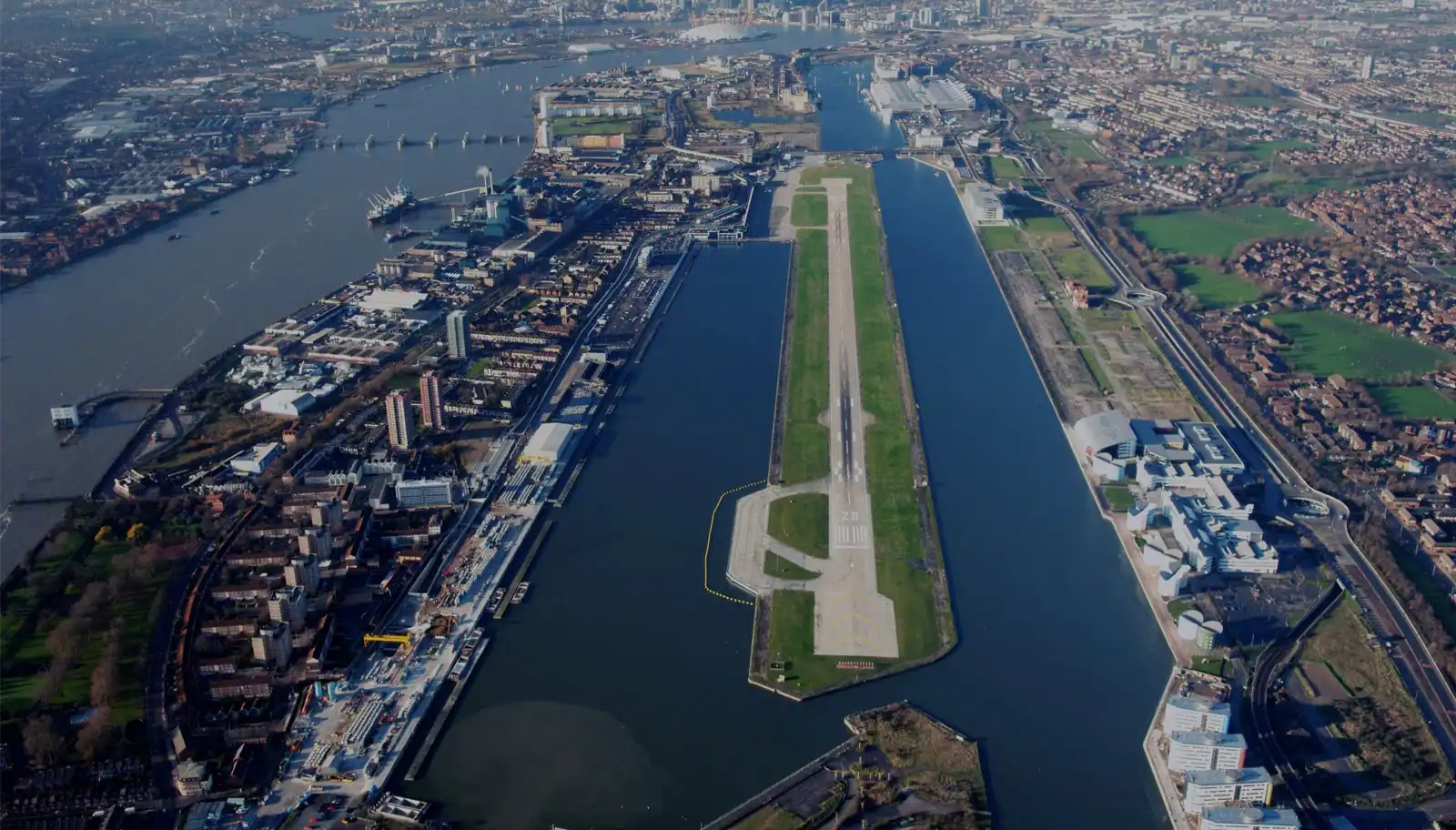 Aerial view of London City Airport with its runway surrounded by Royal Docks and Thames River, showcasing East London's urban landscape