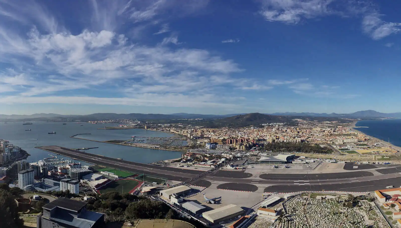 Panoramic aerial view of Gibraltar Airport with runway extending into the bay, cityscape, Mediterranean coastline, and mountains in background