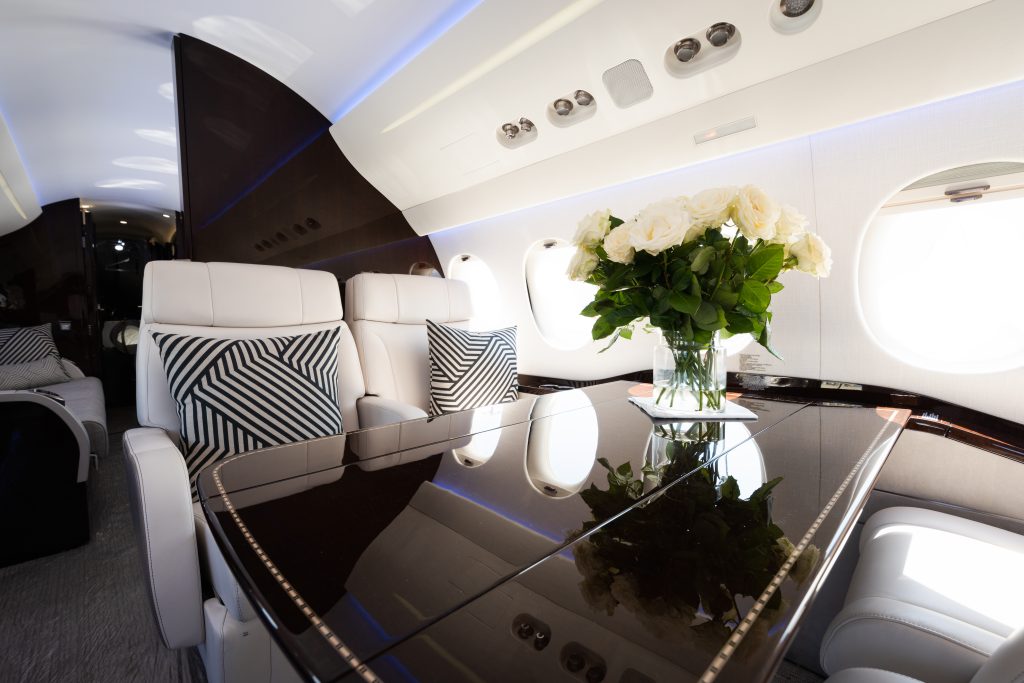 An Interior Shot of a private jet shows a dark wooden table with white flowers next to a set of windows. The dark table contrasts the white seats and gives a modern feel to the aircraft.