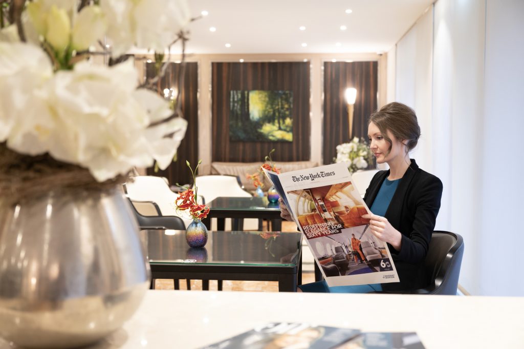 In a VIP airport lounge, a woman sits comfortably, immersed in reading The New York Times. She exudes an air of sophistication, surrounded by a stylish and exclusive atmosphere.
