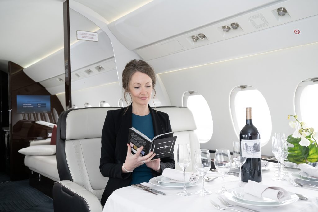 In the private jet's elegant interior, a table set with white orchids, wine glasses, and plates exudes sophistication. A red-haired woman relaxes at the table, engrossed in a book amid the luxurious ambiance.