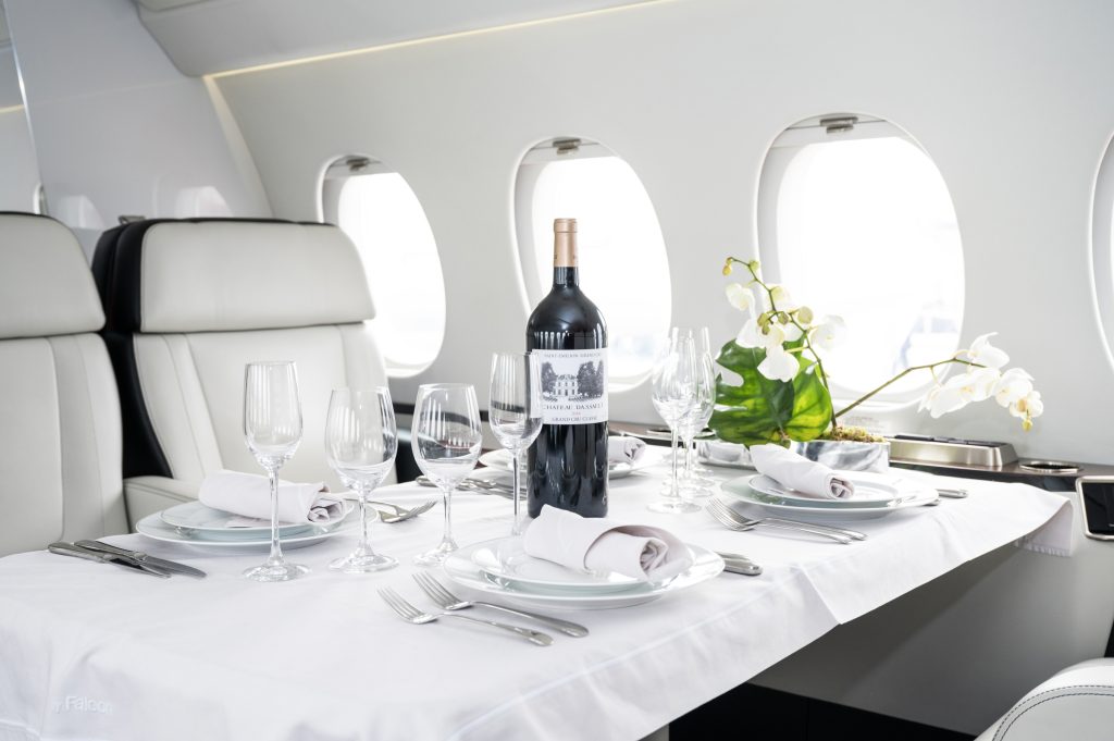 An Interior shot of a private Jet, next to a few windows is a table, adorned with a white tablecloth, wine glasses and 4 sets of plates and cutlery have been laid out in front of the seats, there are also white orchids on the table along with a bottle of wine. 